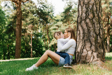 Naklejka na ściany i meble Mom and son love in the park in summer, sitting near a tree