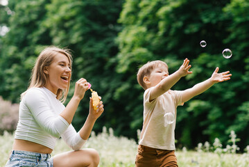 Young Caucasian mother and her happy little son are having fun playing with soap bubbles in the park