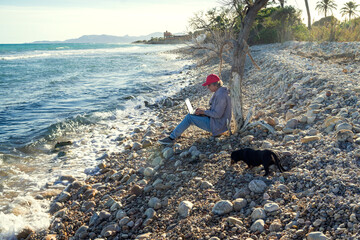 A 50-year-old man is sitting under a tree by the seashore. His loyal dachshund rests beside him. The man is focused on his laptop, which rests on his lap as he works amidst the tranquility of this des