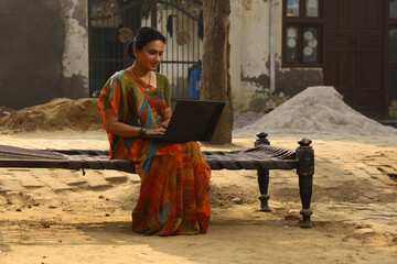 Happy Indian Rural woman sitting on cot outside her home in front yard. In Beautiful saree. Working on a laptop.