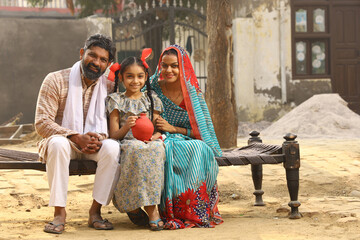 Happy rural Indian family sitting together, daughter is holding a piggy bank in her hand, putting a coin in it, showing the concept of saving money
