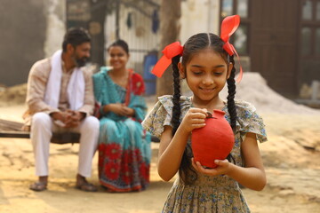 Happy rural Indian family sitting together, daughter is holding a piggy bank in her hand, putting a...