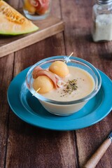 Cold melon soup with thin ham and fresh melon balls in a transparent glass bowl on a brown wooden background. Spanish cuisine.