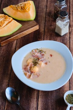 Cold melon soup with thin ham and fresh melon balls in a white ceramic plate on a brown wooden background. Spanish cuisine.