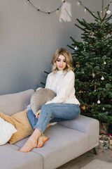 Beautiful young woman is sitting on a cozy sofa with pillows in a room decorated for the new year or Christmas