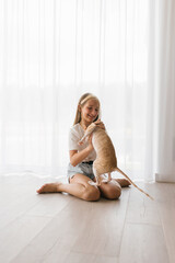 Caucasian teen girl plays with cornish rex cat at home on a sunny day, hugs him and laughs while sitting on the floor in the living room