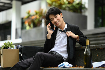 Drunk businessman in suit and tie talking on mobile phone while sitting on stairs with box of stuff. Job loss, unemployed concept
