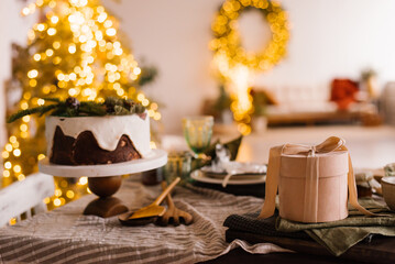 Festive Christmas cake on a wooden stand on a set dining table and a round gift box on a background of lights
