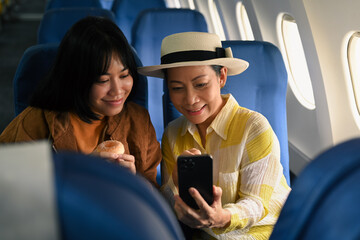 Happy middle aged woman and daughter sitting in passenger airplane and taking picture, waiting for...