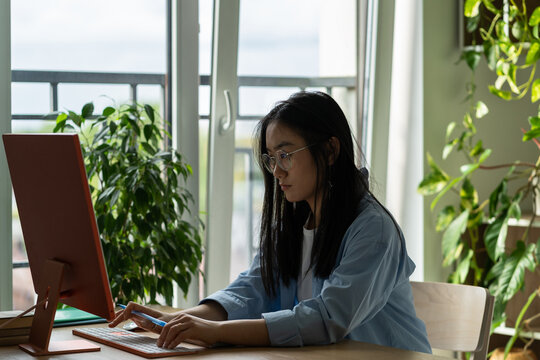 Concentrated Asian woman internet marketer sits at table with computer typing email. Focused Korean girl engaged in SMM promotion of companies in home office near window and houseplant. Work distant