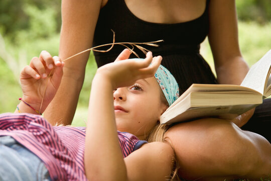 Young girl playing with blade of grass resting head on mother lap

