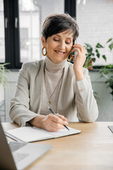 smiling middle aged businesswoman talking on smartphone and writing in notebook in modern office