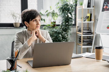 thoughtful businesswoman sitting near laptop and looking away in modern office, executive, ambitious