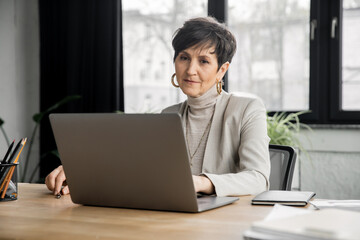 stylish middle aged businesswoman working on laptop in contemporary office space