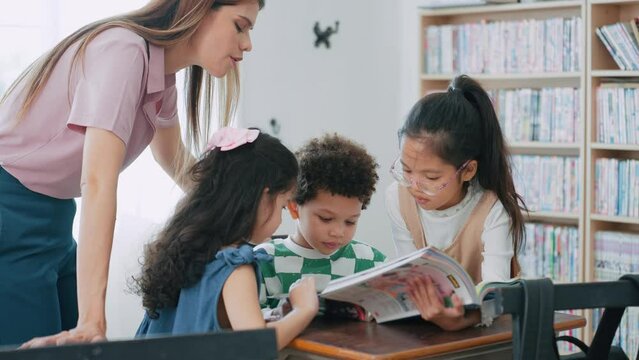 Kindergarten teacher women teaching group of little children reading books in classroom at school. School Kids learning reading a books together with teacher. Education and learning concept