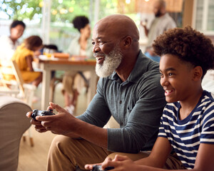 Grandfather And Grandson Sitting On Sofa At Home Playing Video Game Together