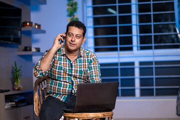 Technology, remote job and lifestyle concept - happy Indian man Talking on phone while working on laptop computer at home office.