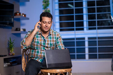 Technology, remote job and lifestyle concept - happy Indian man Talking on phone while working on laptop computer at home office.