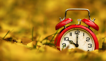Retro red alarm clock in yellow gold autumn leaves. Daylight savings time, fall, morning background with copy space.