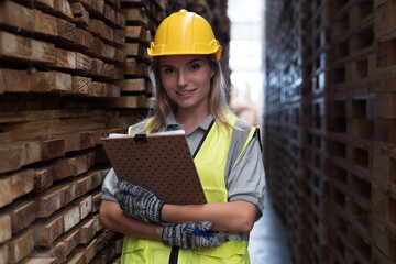 Portrait of female warehouse worker wearing safety uniform holding clipboard and working in wooden warehouse storage. Female worker standing and smiling in wooden warehouse