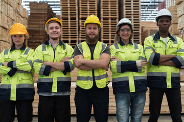 Group of male and female warehouse workers standing together with crossed arms and smiling in industry factory, wearing safety uniform and helmet. Warehouse workers working in warehouse storage