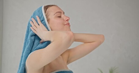 Attractive woman wrapped in towel drying blonde hair with towel after shower. Side view of relaxed caucasian girl wiping wet hair in bathroom, in motion. Concept of hair care routine.