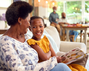 Grandmother And Granddaughter Reading Book On Sofa At Home With Multi-Generation Family Behind