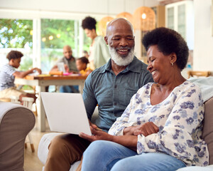 Senior Couple At Home Looking At Laptop Together With Multi-Generation Family In Background