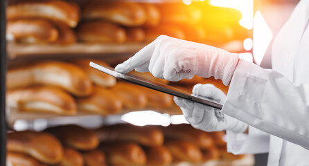 Baker use tablet computer for control quality of craft bread in bakery factory, sun light. Modern...