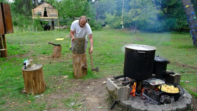 Man with ponytail using axe to try split in half log or stump making firewood for his camp fire while cooking food.