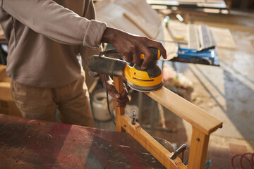 Close up of young black man crafting wooden furniture using electric sanding tool in sunlight, copy space