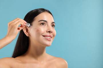Beautiful young woman applying serum onto her face on light blue background. Space for text