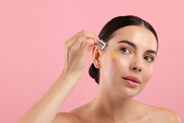 Fototapeta premium Beautiful young woman applying serum onto her face on pink background