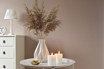 Pampas grass in a white textured vase on a white, round table, next to burning candles.  Delicate nude colors.