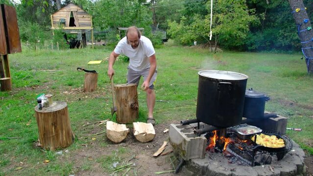 Man with ponytail using axe to split in half log or stump making firewood for his camp fire while cooking food.