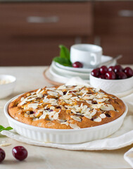 Pie with cherries and almond petals on the kitchen table, in the background a saucer and a cup, a bowl with cherries.