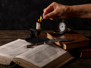 A female hand lights a candle on a table with old books, a quill pen and an antique pocket watch