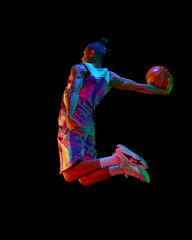Athletic young guy, basketball player in motion during match, jumping with ball against black studio background in neon light. Concept of professional sport, competition, hobby, game, competition