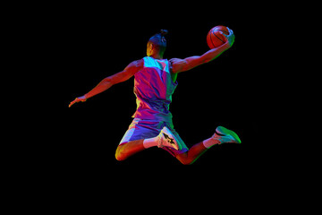 Back view image of young guy, basketball player in sportswear training, jumping and throwing ball against black studio background in neon light. Concept of sport, competition, hobby, game, competition © master1305