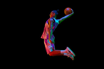 Athletic young guy, basketball player in motion during match, jumping with ball against black...
