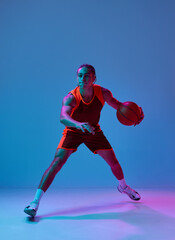 Dribbling ball . Dynamic image of teen boy in uniform, playing basketball against blue studio...