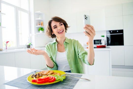 Photo of excited shiny young girl wear green shirt eating breakfast recording video modern gadget indoors kitchen room