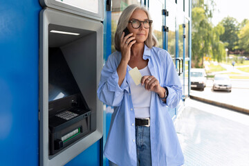 mature adult woman talking on the phone while standing near the ATM