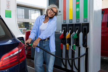 a mature woman with gray hair stands at a gas station and refuels the car for a trip and speaks on...