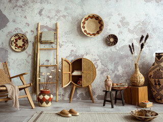 Creative composition of ethnic living room interior with colorful baskets, round rattan sideboard, ladder, stylish armchair, beige plaid, carpet, sculpture, concrete wall. Home decor. Template.