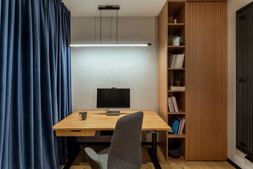 Interior design of office space with wooden desk, stylish gray armchair, navy curtain, modern lamp, computer, wooden bookcase, books, cup and personal accessories. Home decor. Template.