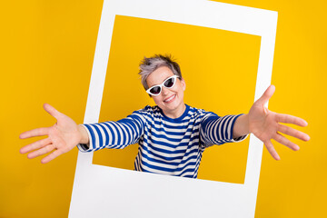 Photo of cool blogger woman pensioner wear stylish sunglass inside paper window zone shooting welcome isolated on yellow color background