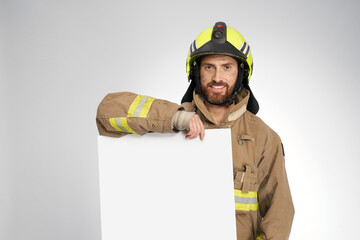 Smiling firefighter in protective helmet holding blank banner in studio. Portrait of bearded man in...