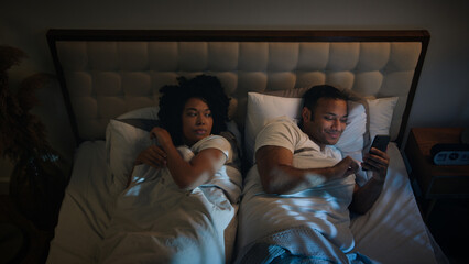 African American couple family in bed bedroom at night angry frustrated woman wife trying sleep offended on husband man gadget addict chatting with phone laughing mobile smartphone addiction problem