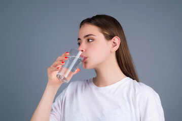 Young woman drinking a fresh glass of water, isolated on studio background. Thirsty woman holding glass drinks still water preventing dehydration, water balance, healthy lifestyle.